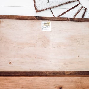 K&ouml;nnte beinhalten: Ein rechteckiges Holzschild mit einer hellen Holzmitte und einem dunkelbraunen Rahmen. Ein kleiner Aufkleber ist oben in der Mitte sichtbar. Das Schild liegt auf einer Holzoberfl&auml;che, mit einer dekorativen Windm&uuml;hle im Hintergrund.