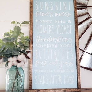以下が含まれることがあります： 「SUNSHINE farmer's markets honey bees & FLOWERS PLEASE thunderstorms chirping birds BUTTERFLIES fresh cut grass picnics in the park & STARS AFTER DARK」と白文字で書かれた薄い青色の木製看板です。