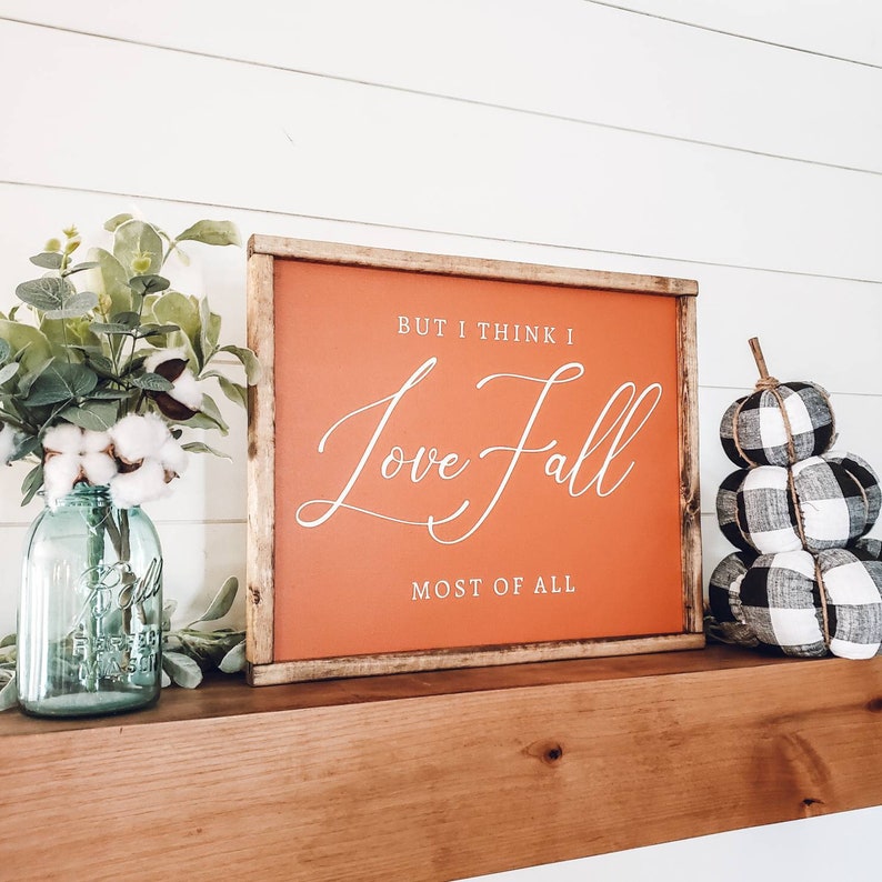 May include: A framed sign with the text "But I think I Love Fall Most of All" in white script on an orange background. The sign is displayed on a wooden shelf with a blue mason jar vase and stacked fabric pumpkins.