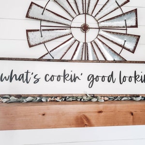 May include: A white wooden sign with a brown frame, hanging on a wooden mantel. The sign reads "what's cookin' good lookin'".