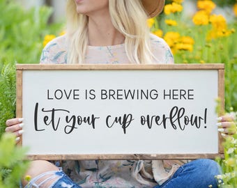 May include: A white wooden sign with black text that reads "Love is brewing here Let your cup overflow!" The sign is being held by a person wearing a floral shirt and blue jeans.