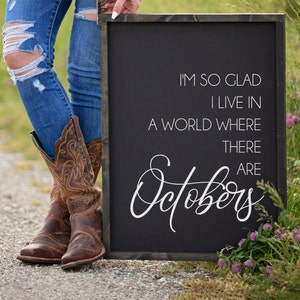 K&ouml;nnte beinhalten: Ein gerahmtes schwarzes Holzschild mit wei&szlig;em Text: &bdquo;I'M SO GLAD I LIVE IN A WORLD WHERE THERE ARE Octobers&ldquo;. Die Person tr&auml;gt Jeans und braune Cowboy-Stiefel.