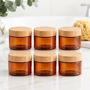May include: A set of six cylindrical amber glass jars arranged in two rows of three. Each jar features a smooth, light-colored bamboo lid that sits flush with the top. The glass is translucent and brown, providing a clear view of the interior.
