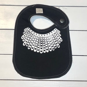 May include: Black baby bib with a white design of a collar with dots. The bib has a white label with the word "Snuggle" on it.