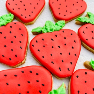 May include: Close-up of several strawberry-shaped cookies. Each cookie has a red icing base with black seed details and a green leaf accent. The cookies are arranged on a white surface, showcasing their vibrant colors and detailed design.