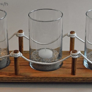 Pode incluir: Um porta-velas de madeira com três porta-velas de vidro. Cada porta-velas de vidro está cheio de areia e uma vela branca. O porta-velas é decorado com corda branca e postes de madeira.