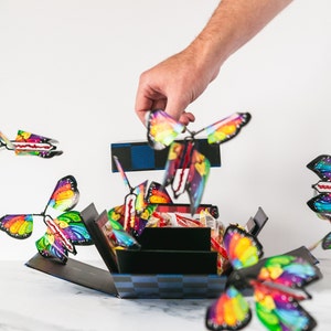 May include: A black and blue checkered box with a variety of colorful butterfly toys popping out of it. The box is open and the butterflies are flying out. A hand is reaching into the box to grab a butterfly.
