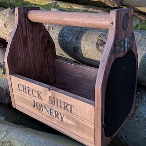 May include: A dark brown wooden caddy with a handle and a chalkboard on one side. The front of the caddy is printed with the words "CHECK SHIRT JOINERY". The caddy is designed to hold items and is made of wood.