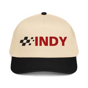 以下が含まれることがあります： ベージュ色の野球帽、つばは黒色。 赤色の「INDY」の文字と、その左側に白と黒のチェッカーフラッグのデザインが施されています。 帽子のてっぺんには黒いボタンが付いています。