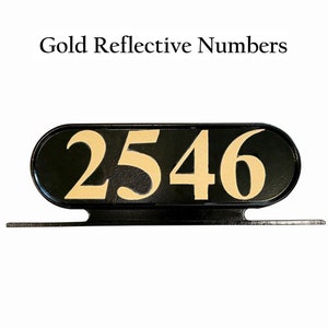 Op de afbeelding: Zwart ovaalvormig huisnummerbord met gouden reflecterende cijfers 2546. Het bord is gemonteerd op een zwarte metalen beugel.