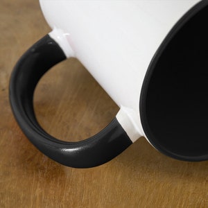 May include: A white ceramic mug with a black handle and a black interior. The mug is sitting on a wooden surface.