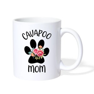 May include: White ceramic mug with a black paw print design, featuring the text "CAVAPOO MOM" in black. The paw print is decorated with pink and orange flowers and green leaves. The mug has a curved handle.