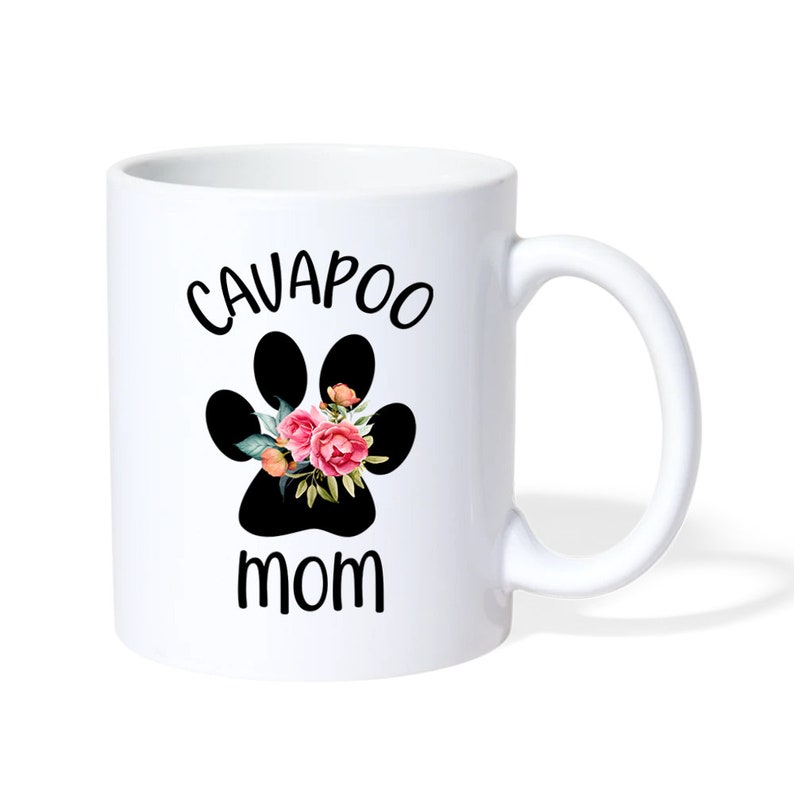 May include: White ceramic mug with a black paw print design, featuring the text "CAVAPOO MOM" in black. The paw print is decorated with pink and orange flowers and green leaves. The mug has a curved handle.