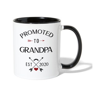 May include: White ceramic coffee mug with a black rim and handle. The mug has black text that reads "Promoted to Grandpa Est 2020" with a red arrow and heart.