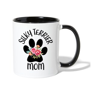 May include: White coffee mug with a black rim and handle. The mug has a black paw print with pink flowers and the text "Silky Terrier Mom" in black.