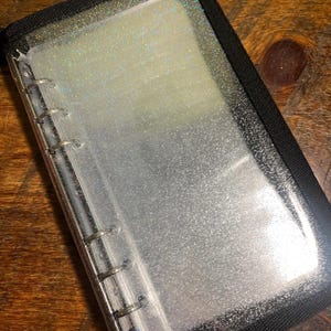 May include: A clear, zippered binder with silver metal rings and a black border. The binder's interior is filled with clear, glittery pages. The binder is closed and sits on a wooden surface.