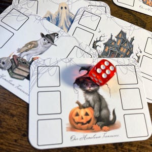 May include: A set of white rectangular game cards featuring Halloween illustrations including a ghost, a haunted house, an owl in a hat, and a kitten in a hat sitting by a pumpkin. A red six-sided die rests on one card. Each card has empty boxes.