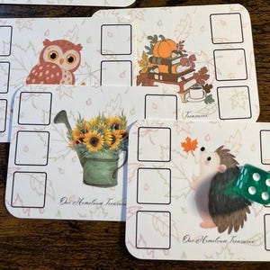 May include: Four rectangular game cards with rounded corners featuring autumn-themed illustrations including an owl, a stack of books with a pumpkin, a sunflower-filled watering can, and a hedgehog. Each card has empty squares and the text Our Hometown Treasures. A translucent green six-sided die rests on the hedgehog card.