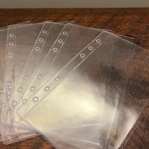 May include: A stack of clear, transparent plastic binder sleeves. Each sleeve has three holes along the top edge for insertion into a binder. The sleeves are arranged in a fan shape, with the top edges visible.
