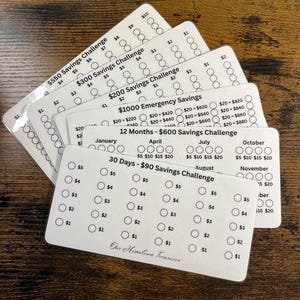 May include: A stack of white savings challenge cards with various amounts and timeframes. Each card features circles to track savings progress. Text includes "$550 Savings Challenge," "$300 Savings Challenge," and "$200 Savings Challenge."