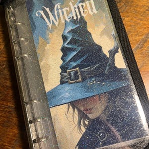 Könnte beinhalten: Ein schwarzer Ordner mit Reißverschluss und einer transparenten Hülle, auf der das Wort "Wicked" und eine Illustration eines Hexenhuts abgebildet sind. Das Bild hat eine blaue und silberne Farbgebung mit Glitzerakzenten.