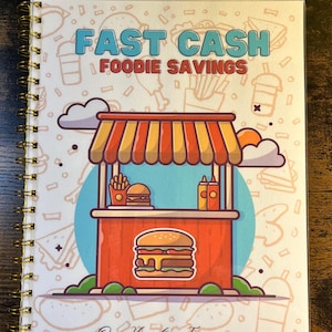 May include: A spiral-bound notebook with a white cover featuring the text FAST CASH FOODIE SAVINGS and Our Hometown Treasures. The central illustration depicts a red food stand with a striped awning, a burger, fries, and condiments.