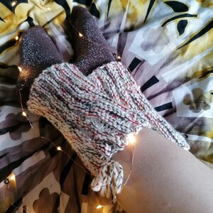May include: A pair of brown socks with silver glitter are tucked into a white and red knitted leg warmer with a long tassel. The leg warmer and socks are on a bed with a patterned bedspread and string lights.