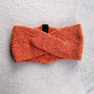 May include: A coral-colored knitted headband with a black fabric accent. The headband is folded in half and has a textured knit pattern.