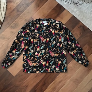 Puede incluir: Blusa negra con estampado de pinceles, cuerdas y borlas en dorado, rojo, azul y blanco. La blusa de manga larga tiene botones en la parte delantera y un cuello clásico. Prenda de estilo vintage.