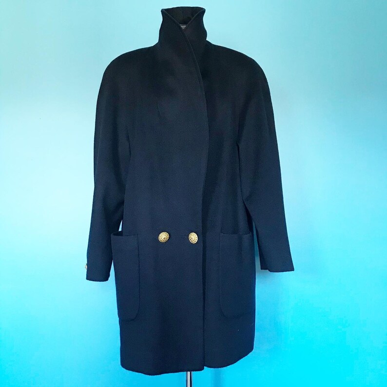 Pu&ograve; includere: Un lungo cappotto blu scuro con collo alto e due bottoni dorati. Il cappotto ha due grandi tasche sul davanti.