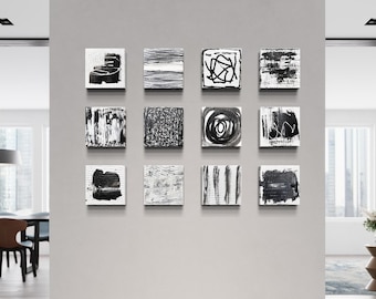 May include: A gallery wall display of 12 black and white abstract paintings in square frames. The paintings are arranged in a grid pattern on a white wall.