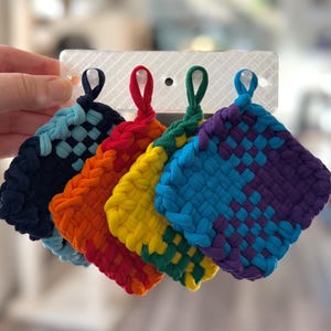 May include: A collection of five colorful, woven pot holders. Each pot holder features a square shape with a loop for hanging. The colors include navy blue, orange, yellow, green, and a blue and purple combination.