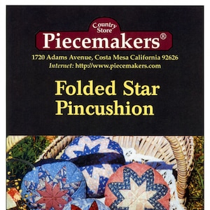 May include: Five folded star pincushions in a wicker basket. The pincushions are made of fabric in various colors and patterns, including blue, pink, red, and white. The pincushions are arranged in a circle, with the basket in the background. The text "Folded Star Pincushion" is printed on the front of the image.