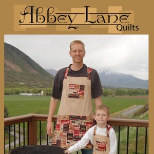 May include: Two people wearing matching brown and red patchwork aprons with the text "BBQ" on them. The aprons have a striped pattern and a pocket. The people are standing on a wooden deck with a black grill in the foreground. The text "Sloppy Joes" is written in a decorative font below the image. The text "AbbeyLaneQuilts.com" and "#217" are written below the text "Sloppy Joes".