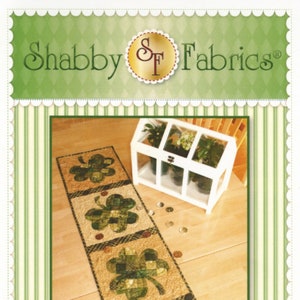 May include: A green and gold patchwork shamrock table runner with a "Patchwork Shamrock Table Runner" title. The runner is made of fabric squares sewn together to create a shamrock design. The runner is on a wooden floor with a small white greenhouse in the background.
