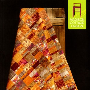 May include: A quilt pattern called "Persimmon Bars" by Madison Cottage Design. The quilt is made of fabric squares in shades of orange, brown, and burgundy. The quilt is hanging on a wooden door.