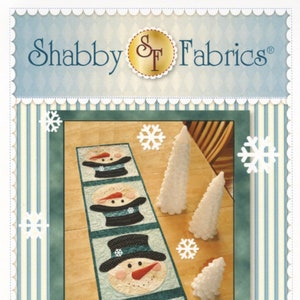 May include: A patchwork snowman table runner with four snowmen in black top hats and a blue background. The table runner is on a wooden table with white snowflake decorations and white pine trees.