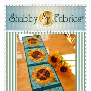 May include: Patchwork Sunflower Table Runner pattern by Shabby Fabrics. The pattern features a table runner with four sunflowers in a row on a blue background. The runner is on a wooden table with a bouquet of sunflowers.