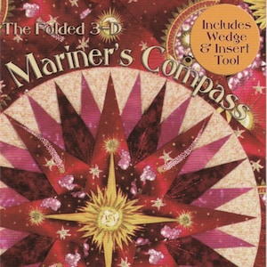 May include: A red, pink, and gold fabric quilt block with a star design. The quilt block is titled "The Folded 3-D Mariner's Compass" and includes the text "Includes Wedge & Insert Tool". The quilt block is signed "Cheryl Phillips".