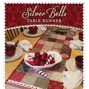 May include: A festive table runner with the text "Silver Bells TABLE RUNNER." The runner features a patchwork design in red, beige, and brown tones. Decorative bells and snowflakes adorn the bottom. A bowl of ornaments sits on the table.