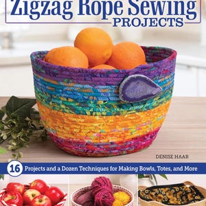 May include: A colorful, handmade rope bowl filled with oranges sits on a wooden surface. The bowl is made with a zigzag sewing technique, featuring vibrant hues of purple, blue, green, yellow, and orange. The book cover reads "Creative Zigzag Rope Sewing Projects."