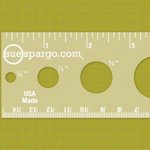 May include: A white plastic ruler with a series of circles cut out. The circles are labeled with measurements in inches: 1/4", 1/2", 3/4", 1", and 1 1/4". The ruler also has a scale in inches from 1 to 5. The ruler is labeled "suelspargo.com" and "USA Made".