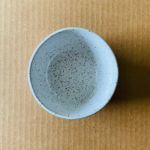 May include: A white ceramic bowl with a speckled gray glaze. The bowl is empty and sitting on a brown surface.