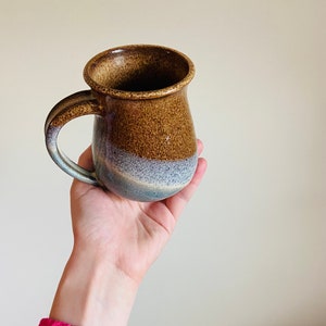 May include: A handmade ceramic mug with a brown and blue glaze. The mug has a rounded body, a sturdy handle, and a slightly flared rim. The color transitions from brown to blue, creating a unique and rustic appearance. Perfect for coffee or tea.