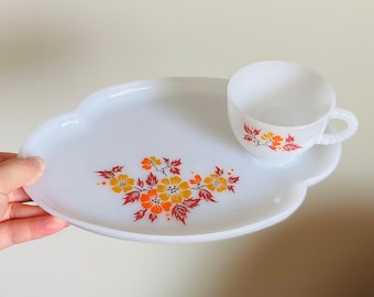 May include: White ceramic snack plate with a scalloped edge and a floral design in shades of orange, yellow, and brown. A matching white ceramic cup with a floral design is on the plate.
