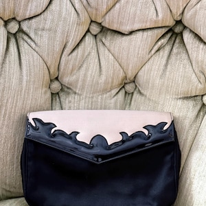 May include: A black satin clutch with a light pink flap. The flap is trimmed with a glossy, dark blue, flame-like design. The bag is set against a tufted, neutral-toned background.