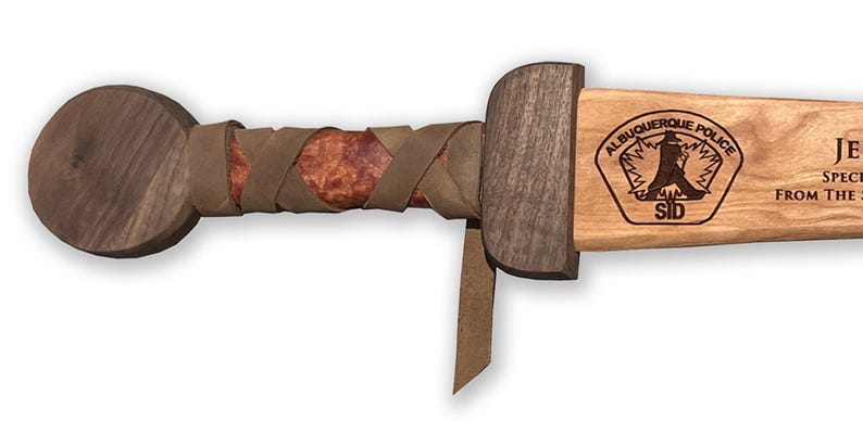 May include: A wooden sword with a dark brown handle and a light brown blade. The handle is wrapped in brown leather. The blade has the Albuquerque Police logo and text.