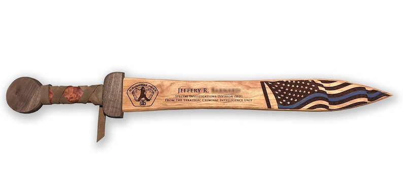 May include: A wooden sword with a round pommel and a wrapped handle. The blade features an American flag design with a blue line. Text includes a name, police unit, and division.