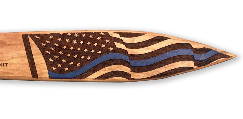 May include: Wooden paddle with a carved American flag design. The flag features stars, stripes, and a blue line. The paddle is made of light-colored wood with a dark wood flag design.