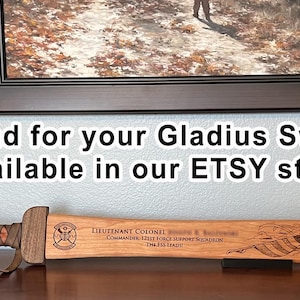May include: A wooden Gladius sword with a brown handle and a light brown blade, resting on a dark surface. The blade has engraved text and an American flag design. The text reads "Stand for your Gladius Sword available in our ETSY store."